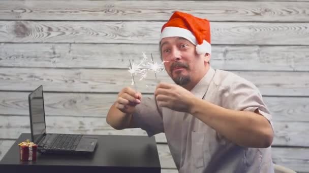 Knappe man met Santa symbool is met behulp van een laptop en verheugt zich met sparkler vuur — Stockvideo