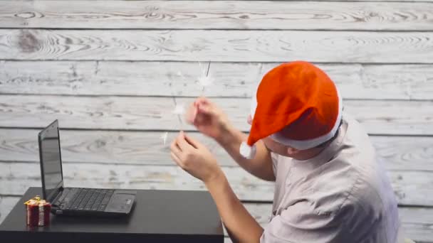 Knappe man met Santa symbool is met behulp van een laptop en verheugt zich met sparkler vuur — Stockvideo