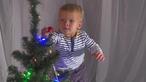 Dwuletni chłopiec bawi się zabawkami Boże Narodzenie na choinkę, z bliska. Portret dziecka w pobliżu Choinka. — Wideo stockowe