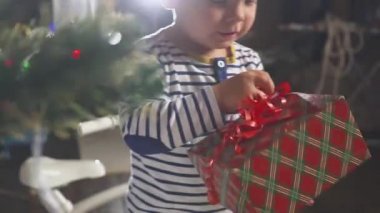 diğer bir sürü hediye kutuları ile bir Noel ağacı arkasında kırmızı kurdele ile bir hediye alma iki yaşındaki küçük tatlı çocuğa. Noel ağacı ışıkları ve altın top ile dekore edilmiştir. Tatil.