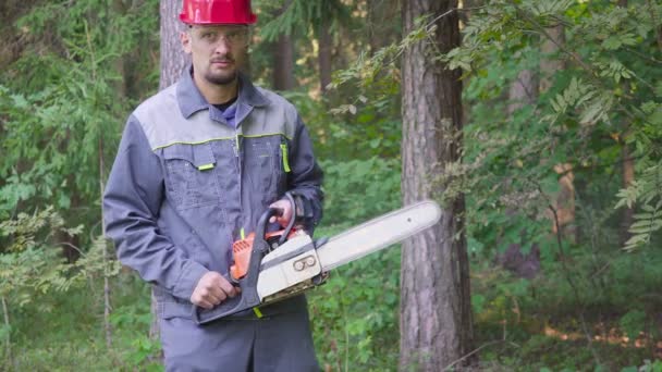 Retrato del trabajador en un casco con una motosierra en el bosque. El hombre mira severamente a la cámara — Vídeos de Stock