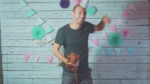 Young attractive man on rocking horse with a cap on his head. Happy birthday concept — Stock Video