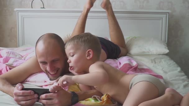 Батько і син, лежачи на ліжку і використання мобільного телефону разом — стокове відео