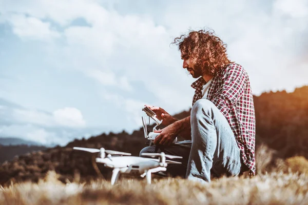 A handsome adult curly man with the beard is programming via remote control the flying route of his quadcopter (in a defocused foreground) before the flight, with hills and sky in a blurred background
