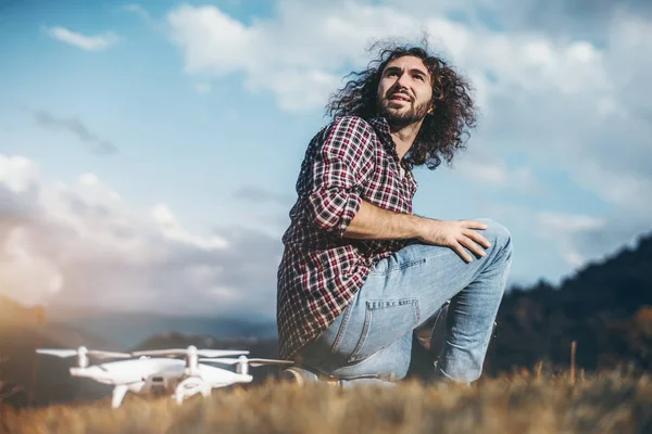 An adult bearded hipster guy with curly hair is looking back while sitting next to his drone on the ground on a sunny day with mountains in a defocused background, prepared to launch his quadcopter