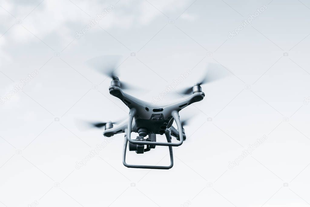 UAV quadcopter with an underslung digital camera is filming an online broadcast; the white flying drone is making test flight, with bright sky in the background