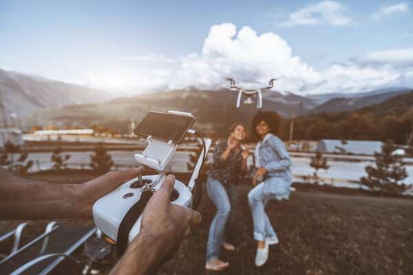 Широкоугольный снимок рук оператора с дистанционным контроллером на переднем плане, управляющим летающим беспилотником перед которым снимаются две девушки разных рас, позирующие для трансляции, горы позади
