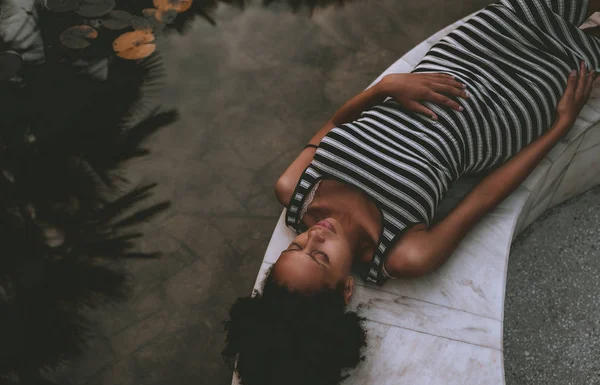 年轻的非洲裔美国孕妇在条纹礼服和卷曲的非洲头发是在夏季黄昏休息的大理石长凳旁边的池塘与睡莲和棕榈树的反射 — 图库照片