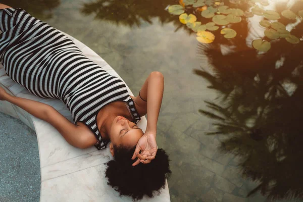 疲倦美丽怀孕非洲裔美国女孩与卷曲的非洲头发是休息和铺设在池塘附近的大理石长凳与睡莲在一个阳光明媚的夏日早晨 棕榈剪影在水中 — 图库照片