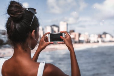 Cityscape ve kıyı şeridi bir fotoğraf çekici smartphone ile Afrikalı-Amerikalı kadın arkadan görünüş; Şehir plaj bir gözlem noktasından çekim Genç bir siyah kız arkadan görünümü