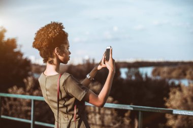 Onu cep telefonundan; bir manzara fotoğrafta şirin Afrikalı-Amerikalı genç kadın arkadan görünümü Dikiz siyah bir kızla cityscape yüksek noktadan smartphone ile fotoğraf çekimi
