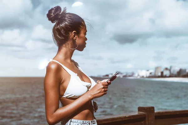 年轻耀眼的巴西女性站在与她的智能手机在观察点的海洋旁边 若有所思地看着距离 而等待消息答复从她的朋友 — 图库照片