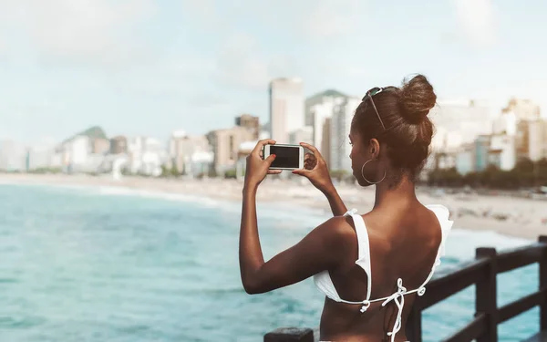 从后面的一个精致的巴西女孩在泳装使用她的手机拍摄的海岸线全景照片 一个年轻苗条的黑色女性的后方视图与智能手机拍摄海滩 — 图库照片