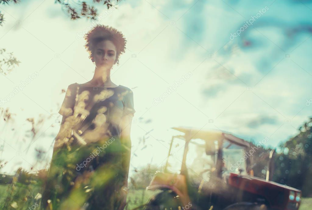 Creative shot of a young serious black girl with curly Afro hair standing on the field with tractor behind; African-American girl outdoors with field engine in the background, shallow depth of field