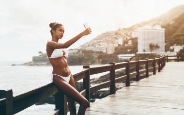 Sevimli siyah kız mayo bir selfie açık havada okyanusun önünde alıyor; genç çekici Brezilyalı erkek kendini ahşap çit yaslanmış iken Smartphone cep telefonu ile fotoğraf çekmek