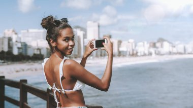 Oynak genç sevimli siyah erkek yarı-kameraya şehir gözlem noktada duran ve onun smartphone ile çekim yaparken cityscape, plaj ve okyanus sahil şeridine dönüm