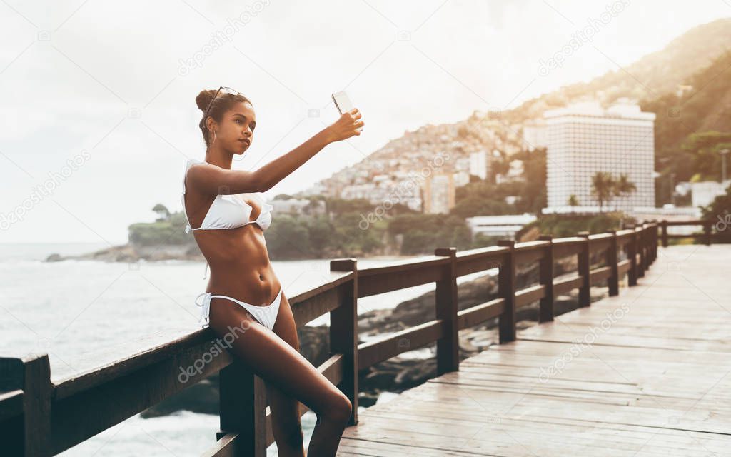 Cute black girl in the swimsuit is taking a selfie outdoors in front of the ocean; young charming Brazilian female is photographing herself on the smartphone while leaning against the wooden fence