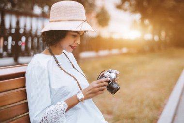 Genç enfes Brezilyalı erkek şapkalı elleri açık havada bankta bir retro film kamera ile; bir vintage fotoğraf makinesi ile mutlu siyah kız parkta tahta bankta oturuyor