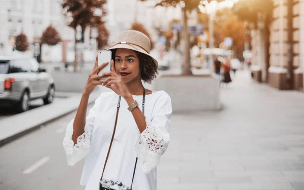 帽子および白いブラウスのかわいい若いアフリカ系アメリカ人観光客の女の子は テキスト メッセージのコピー場所の大通りに立っている間彼女のスマート フォンのカメラを使用して都市の写真を撮っています — ストック写真