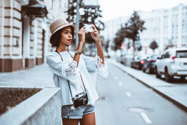 Genç sevimli siyah kız şapkalı sokağın yanında birden fazla otomobillerde ufuk arka plan ve metin mesajı için sağdaki kopyalama alanı alanı ile onun smartphone kullanarak fotoğraf çekmek