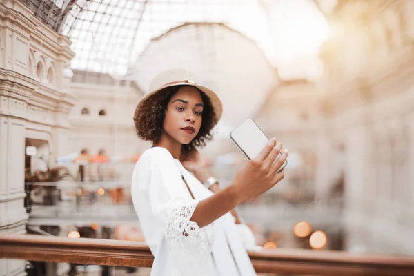 真のチルトシフト セレクティブ フォーカスで撮影 帽子でかわいい若い黒観光客女性は巨大なショッピング モールの屋内で彼女のスマート フォンで Selfie を取っています 手と顔の選択と集中 — ストック写真