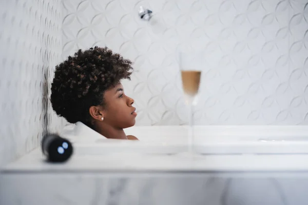 一个疲惫的年轻卷曲的黑色女孩铺设 放松在室内洗澡 听音乐形成一个无线扬声器旁边的她和一杯白葡萄酒在前景的侧面视图 — 图库照片