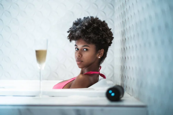 一个迷人的年轻非洲裔美国女孩的肖像在浴室铺设和放松在浴缸与白色的葡萄酒和无线扬声器在前面的弥散前景 — 图库照片