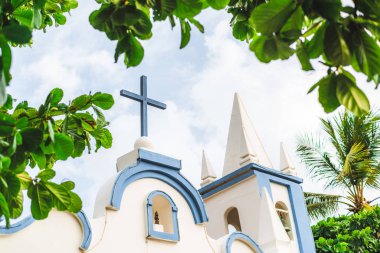The exterior of a small church Saint Francisco de Assis located in state Bahia of Brazil, in the resort Praia do Forte; with blue wooden cross on the top, the chapel with the bell and frame of leaves clipart