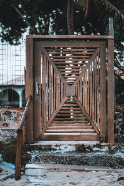 A self-made old wooden door between a street and a yard: multiple strips of wood in rectangular form concentrically converging to the center, several concrete steps and the sand in the foreground