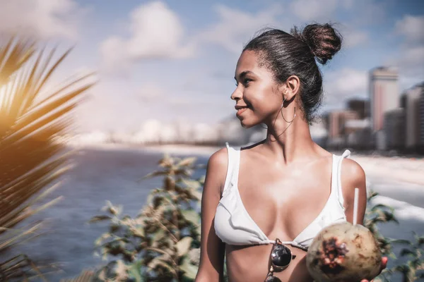 Portrait of young charming Brazilian female standing not far from the coastline with coconut and looking aside, with copy space place for text, your logo or advertising message on the left, sunny day