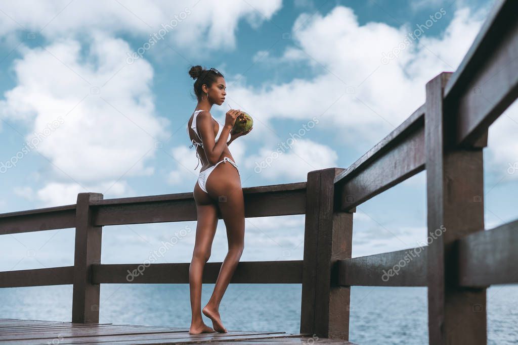 A svelte sexy young black girl in a white swimsuit is thoughtfully looking at the ocean horizon while standing near a wooden fence and drinking coconut water from coco using the the straw