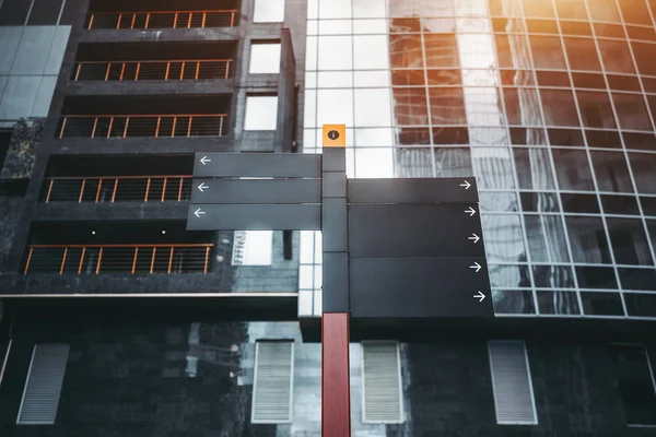 Mock-up of empty urban informational waypoint markers pointing to the left and right made of a black metal; signpost template with a white arrows on it, with an business office building behind