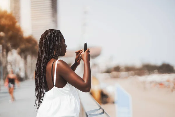 可爱的黑色女孩穿着白色的礼服是拍摄海和海滩使用她的手机相机 一个年轻的非洲女性的侧面看到辫子通过她的智能手机在街上拍照 — 图库照片