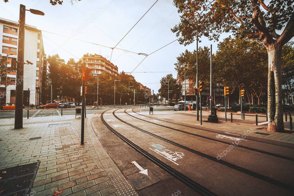 Wide-angle view of an urban autumn street with tramways turning right, multiple cars, traffic lights, trees, road signs on the ground and on poles, residential buildings around, fallen leaves below