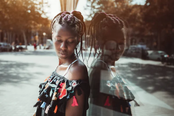 迷人的年轻黑人女性与辫子是靠在玻璃墙上 看着她的肩膀 一个可爱的非洲女孩的肖像编织的头发 在衣服附近的一个巨大的窗口在街上 — 图库照片