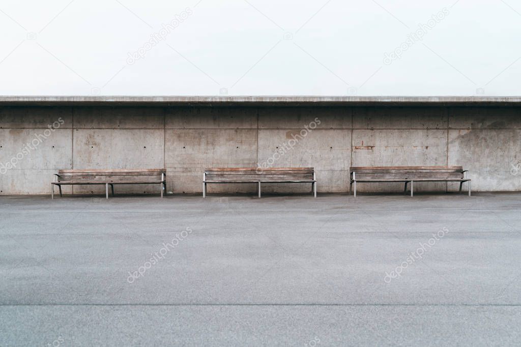 Wide-angle minimalist shot of three wooden benches with metal legs standing in front of a grunge concrete wall with small holes, a huge empty area of the asphalt with a single stripe in the foreground