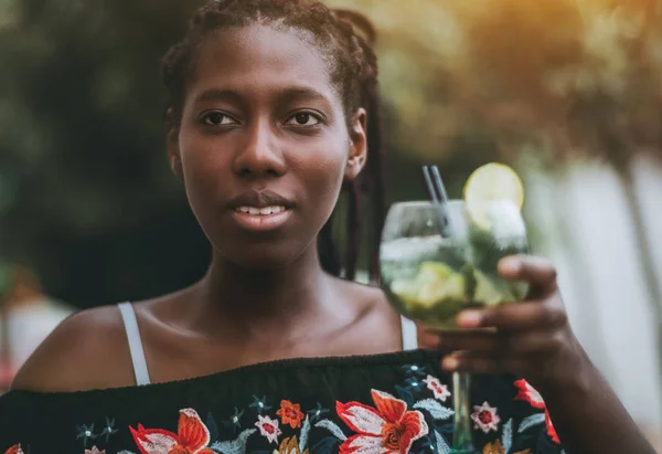 Portrait of beautiful young black female in dress with the flower pattern on it, sitting in an outdoor bar and drinking cocktail; African girl with a glass of delicious mojito in a street cafe