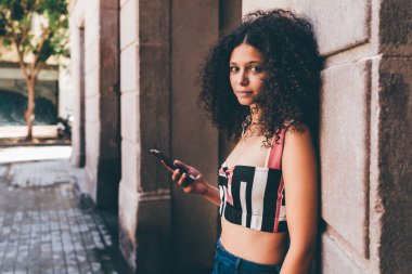 Duvar açık havada karşı yaslanmış ve onun smartphone tutan genç güzel Fas kadın turist portresi; sevimli kıvırcık Afrikalı-Amerikalı kadın sokakta Barselona cep telefonu ile