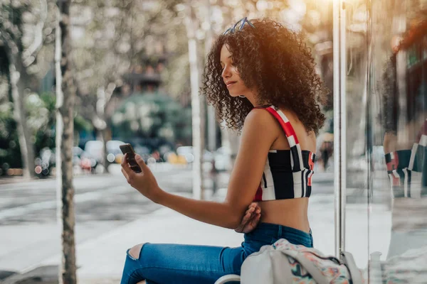 アフリカ系アメリカ人女性を魅力的な内部都市バス停のベンチに座って 彼女スマート フォンを介してビデオ通話携帯電話でアフロ巻き毛の見事な女性がバスを待ってください — ストック写真