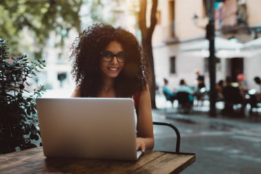 Neşeli genç kadın freelancer laptopunu açık bir kafede çalışıyor; belgili tanımlık netbook sokak çubuk, Barcelona tahta masada otururken kullanarak kıvırcık saçlı beyaz erkek gülümseyen