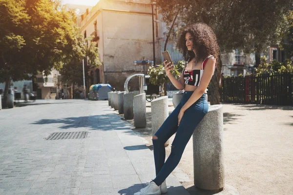 迷人的年轻白种女性与笨重的卷发在一个休闲的衣服是靠在巴塞罗那街头的一个石头杆 并通过智能手机发送信息给她的男朋友 — 图库照片