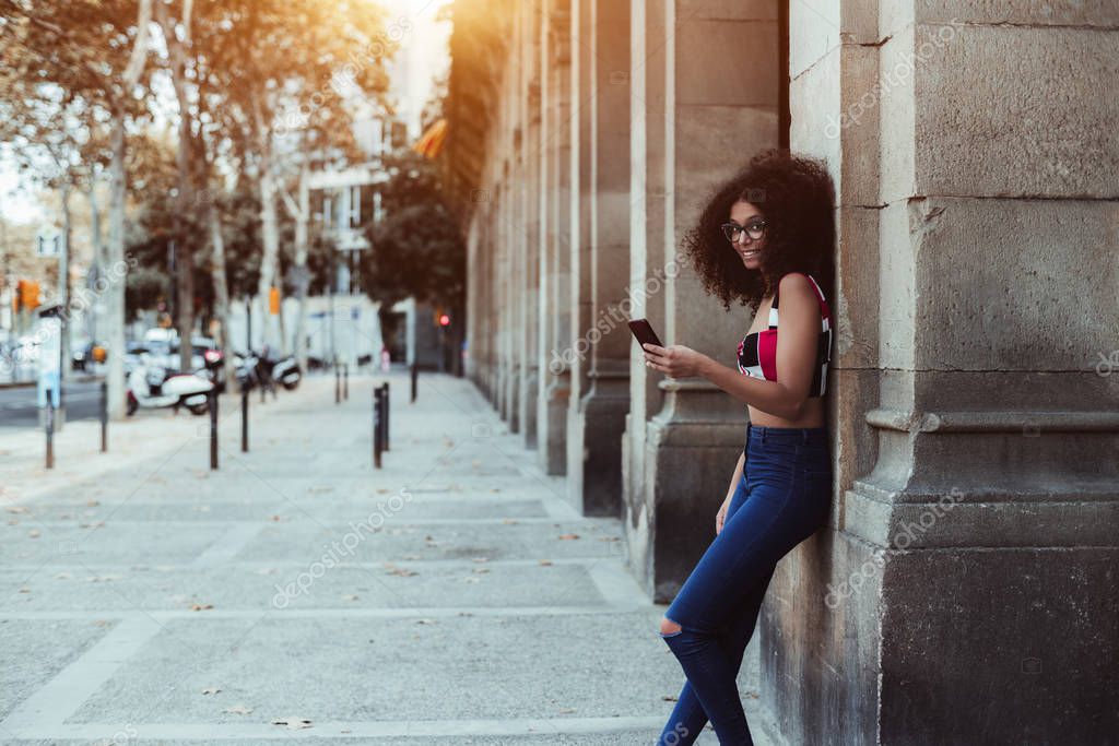Smiling cute girl with curly bulky hair is leaning against a stone pillar outdoors and looking at the camera while holding smartphone in hand, with a copy space area on the left for message or logo