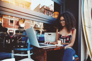 Bir neşeli genç kıvırcık beyaz kadın gözlük içinde laptopunu açık bir kafede çalışıyor; gülümseyen şirin kız bardaklarda netbook sokak bir barda oturmuş ve kahve içme kullanma