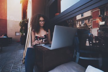 Sevimli genç kadın girişimci kıvırcık afro saçlı ve gözlük bir açık bar bir fincan lezzetli kapuçino holding ve kahve molası sırasında onun projeleri bitirmek için onu laptop kullanma içinde oturuyor