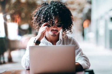 Ciddi kıvırcık Asya adam girişimci yuvarlak güneş gözlüklerini onun dizüstü bilgisayar ve uzaktan onun serbest projeler üzerinde çalışmaya açık bir kafede otururken doğrultma; sığ derinlik-in tarla