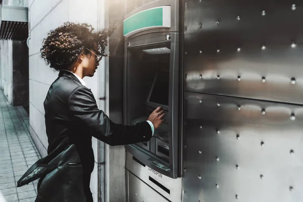 Side view of curly businessman in a formal suit and fancy sunglasses withdrawing money from a bank card using a street cash machine ATM, with a copy space place on the right for your logo or advert