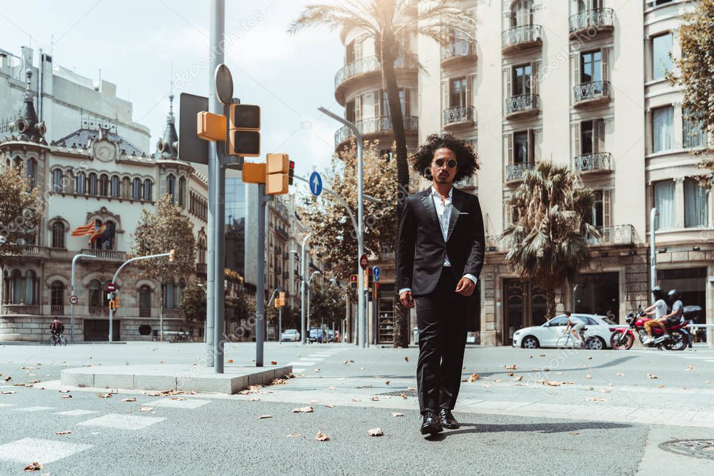 A handsome curly Asian businessman in sunglasses and a formal suit is confidently crossing the road on the crosswalk near traffic lights, with facades of buildings in the background, Barcelona, Spain