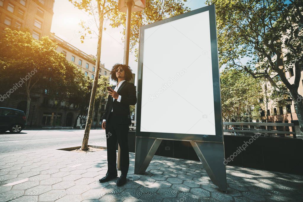 Elegant curly hipster in sunglasses, formal suit, and with a beard is using his smartphone outdoors near a blank white mockup of street information billboard, man entrepreneur near the banner template