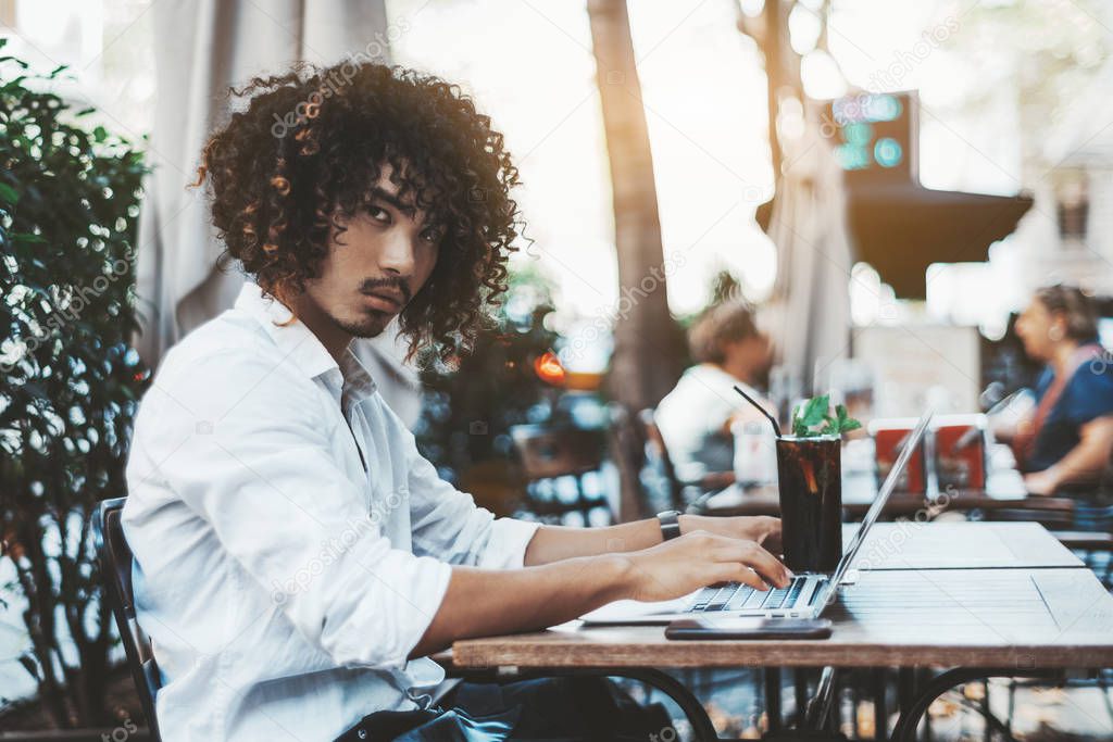 Portrait of a young curly Asian guy in a white shirt using the laptop in an outdoor bar with a glass of a delicious ice tea; a bearded handsome guy in street cafe with a cocktail using the netbook