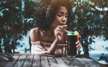 Susuz bir göz kamaştırıcı kıvırcık biracial kadın lezzetli soğuk buzlu çay veya bir nane kadeh kokteyl holding ve yalnız bir sıcak yaz gününde sokak bir barda otururken bir saman kullanarak içme